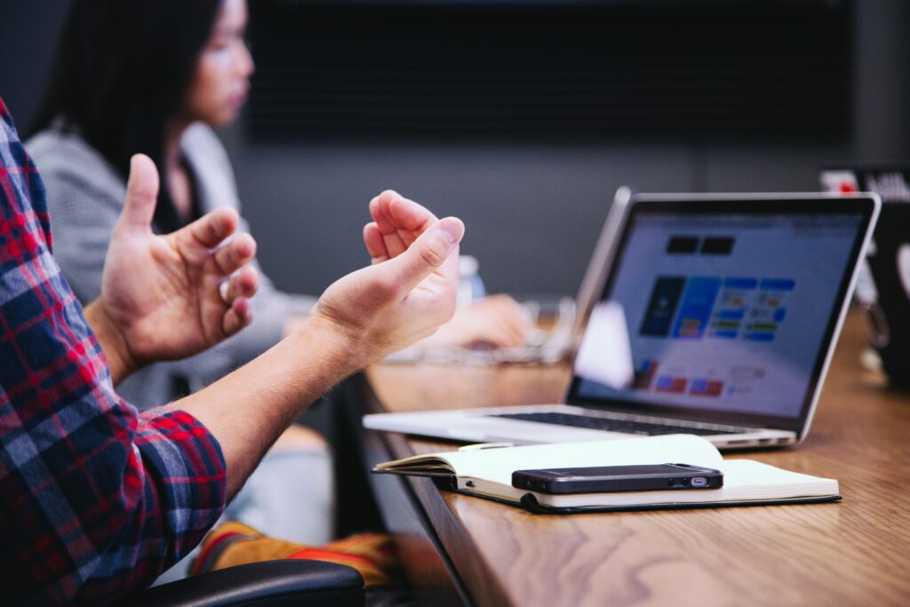 Someone speaking with their hands while sitting at a boardroom table with their laptop in front of then, a blurred woman is sitting in the background | Nextworld