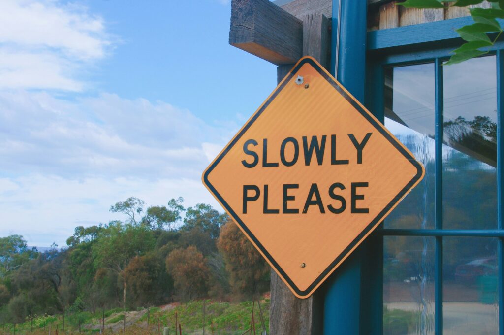 A yellow road sign with the words 'slowly please' across the front of its diamond-shaped face, in the background is a lush forest | Brake Samsara