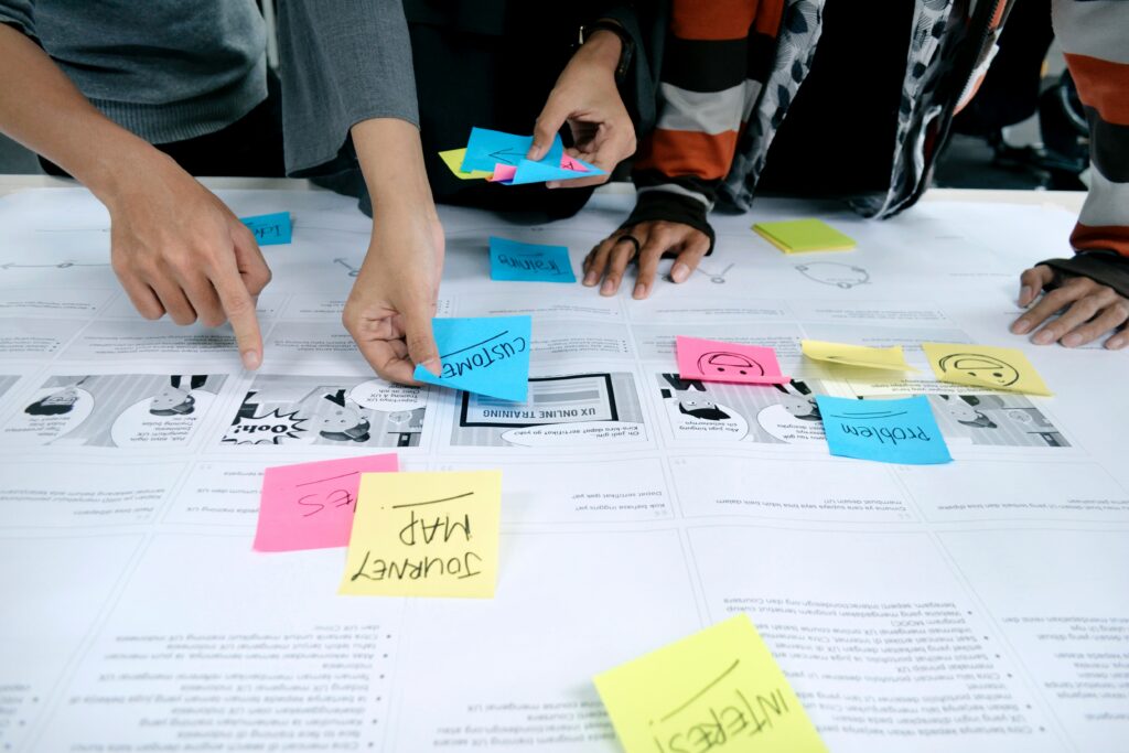 top down view of a cluttered work desk, with multiple sticky notes and a few people standing over the desk, pointing at things | Unit4 research