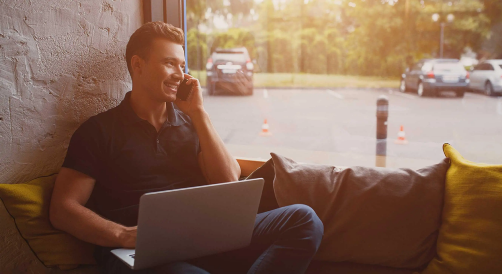 man sitting on next to window with a laptop talking on the phone